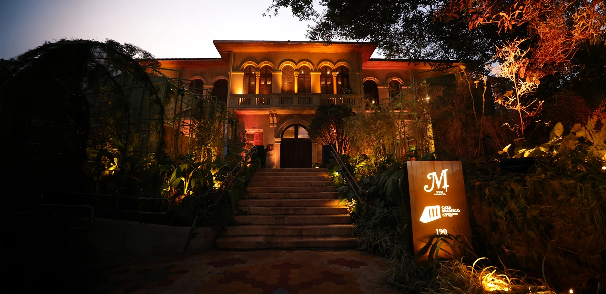 Um grande edifício histórico com janelas em arco e colunas é iluminado ao entardecer, com uma placa iluminada com a letra M perto da entrada, rodeada por plantas exuberantes.