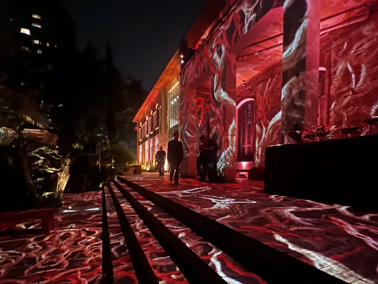 Área externa de um edifício iluminado à noite com projeções de luz em tons de vermelho e branco sobre o chão e as paredes. Algumas pessoas caminham pela escadaria em direção à entrada, enquanto árvores e vegetação aparecem à esquerda, criando um ambiente cênico e artístico.