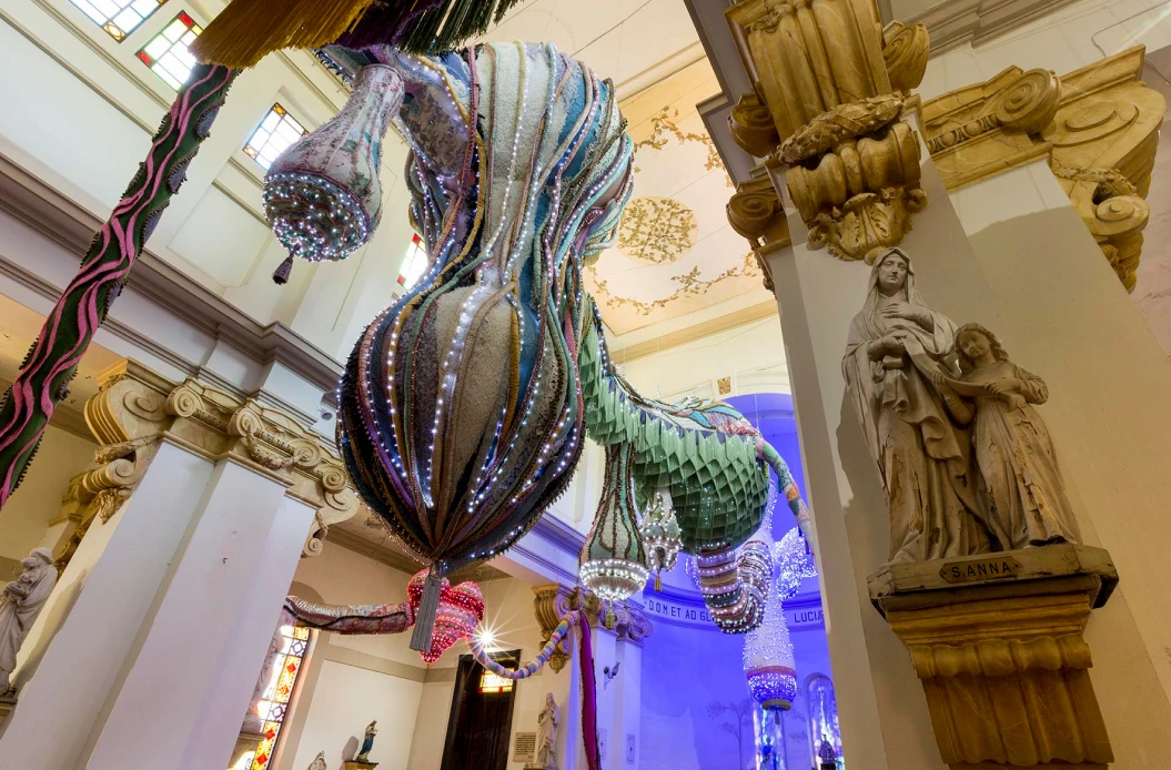 Interior de um edifício histórico com colunas ornamentadas e esculturas religiosas. No centro, uma grande instalação artística contemporânea suspensa, composta por formas orgânicas e alongadas, cobertas por materiais coloridos e luzes brilhantes. Ao fundo, vitrais e paredes decoradas contrastam com a obra moderna pendurada no espaço.