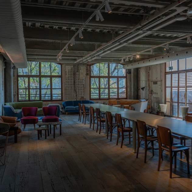 Sala espaçosa em estilo industrial com grandes janelas, mesa comprida com cadeiras de madeira, sofás coloridos, poltronas e luz natural abundante. Aconchegante e convidativa.