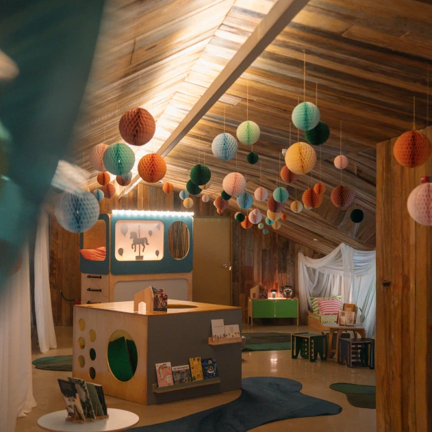 Uma aconchegante sala de jogos infantil com teto de madeira, decorada com lanternas de papel coloridas penduradas. Há livros, brinquedos e uma pequena área de estar.