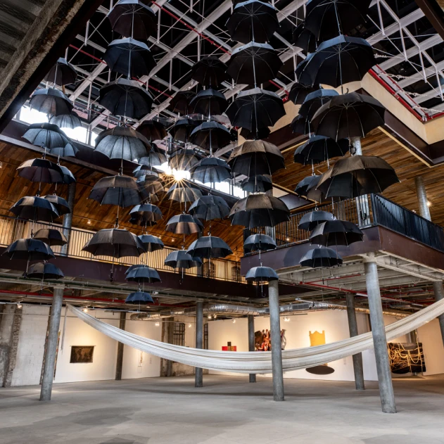 Dezenas de guarda-chuvas pretos estão suspensos em um teto alto de uma galeria de arte, criando um efeito dramático acima de uma grande rede branca esvoaçante.