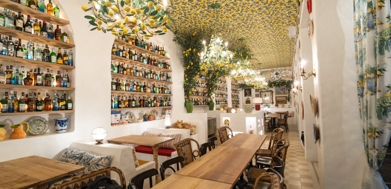 Interior iluminado de um restaurante decorado com tema mediterrâneo, exibindo paredes com prateleiras cheias de garrafas, mesas de madeira, cadeiras de vime e um teto coberto por estampas de limões e folhagens.